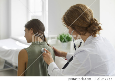 Female doctor with a medical stethoscope examining the lungs of a teenage patient 101047757