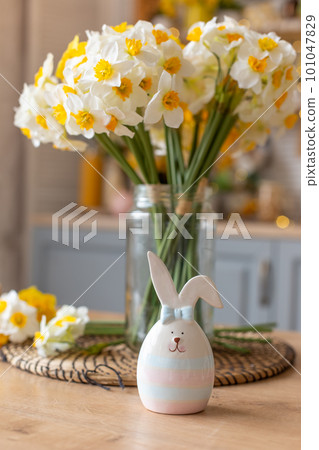 A ceramic figurine of a cute striped hare stands on the kitchen table next to a glass vase with daffodil flowers. 101047829