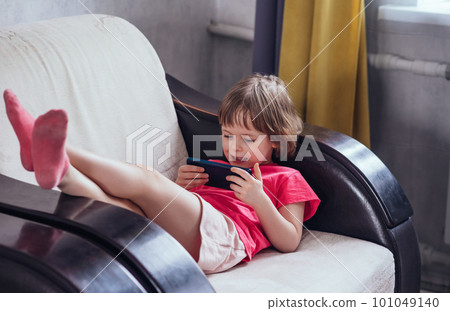 Smiling girl lies in an armchair in a room by the window and and using phone 101049140