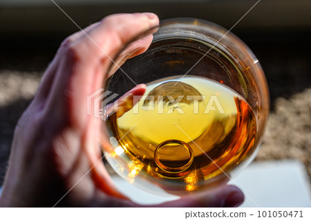 Caucasian hand holding a glass of amber colored liquid with a wedding ring at the bottom 101050471