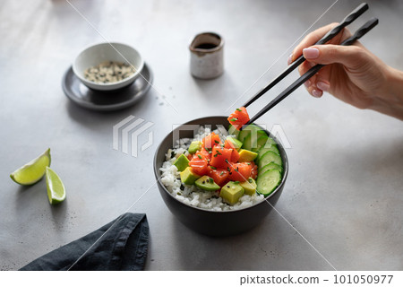 woman's hands holding chopsticks with a slice of salmon and eating a hawaiian poke bowl. 101050977