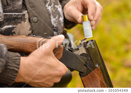Selective focus of the hands of a man in a camouflage protective suit who loads a double-barreled shotgun. The concept of hunting. Selective focus of the hands of a man in a camouflage protective suit who loads a double-barreled shotgun. The concept of hunting. 101054349