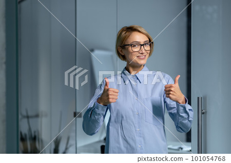 Portrait of a young beautiful female student standing in the office, campus, library. He looks at the camera with a smile, shows his fingers super, success. Portrait of a young beautiful female student standing in the office, campus, library. He looks at the camera with a smile, shows his fingers super, success. 101054768