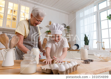 Happy family in kitchen. Grandmother and granddaughter child cook in kitchen together. Grandma teaching kid girl roll out dough bake cookies. Household teamwork helping family generations concept 101054899