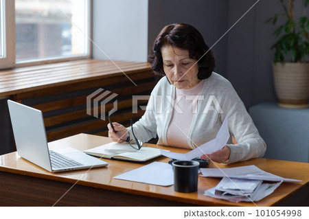 Middle aged senior woman sit with laptop and paper document. Pensive older mature lady reading paper bill pay online at home managing bank finances calculating taxes planning loan debt pension payment Middle aged senior woman sit with laptop and paper document. Pensive older mature lady reading paper bill pay online at home managing bank finances calculating taxes planning loan debt pension payment 101054998