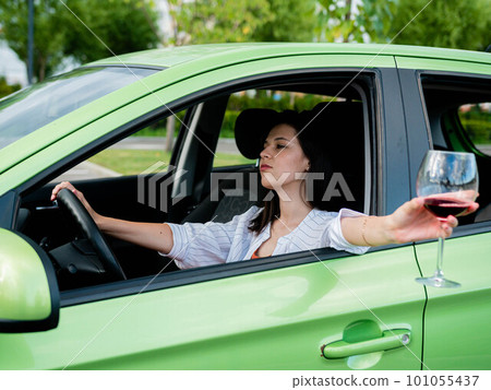 Young beautiful smiling woman driving a car with a glass of wine in her hand. Drinking alcohol while driving. Drunk young girl driving a car Young beautiful smiling woman driving a car with a glass of wine in her hand. Drinking alcohol while driving. Drunk young girl driving a car 101055437