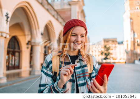 Outdoor shot of pretty beautiful blonde haired young woman uses smartphone technology for writing web review browses website wears spectacles and raincoat carries bag poses Outdoor shot of pretty beautiful blonde haired young woman uses smartphone technology for writing web review browses website wears spectacles and raincoat carries bag poses 101055645