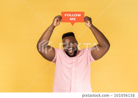 Portrait of cheerful excited bearded young adult man blogger wearing pink shirt standing card with fallow me inscription. Indoor studio shot isolated on yellow background. Portrait of cheerful excited bearded young adult man blogger wearing pink shirt standing card with fallow me inscription. Indoor studio shot isolated on yellow background. 101056138