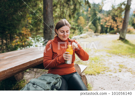 Happy redhead Woman in active trekking clothes having a halt after hiking. Hiker drinking water from water bottle or hot drink from yellow thermos Happy redhead Woman in active trekking clothes having a halt after hiking. Hiker drinking water from water bottle or hot drink from yellow thermos 101056140