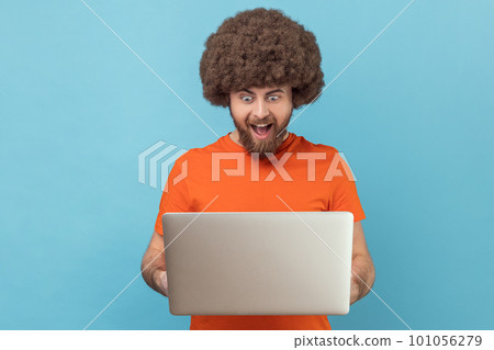 Portrait of excited amazed man with Afro hairstyle wearing orange T-shirt holding notebook and looking at display with excitement, playing computer games Indoor studio shot isolated on blue background Portrait of excited amazed man with Afro hairstyle wearing orange T-shirt holding notebook and looking at display with excitement, playing computer games Indoor studio shot isolated on blue background 101056279