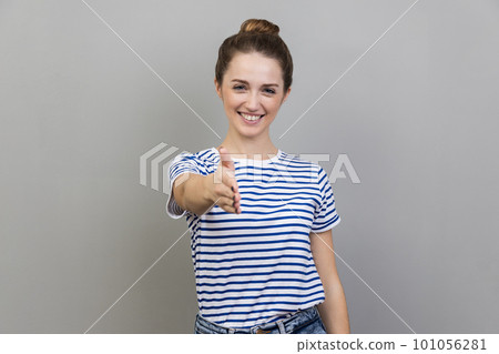 Portrait of smiling friendly woman wearing striped T-shirt holding hand for handshake, happy to make agreement, successful partnership. Indoor studio shot isolated on gray background. Portrait of smiling friendly woman wearing striped T-shirt holding hand for handshake, happy to make agreement, successful partnership. Indoor studio shot isolated on gray background. 101056281
