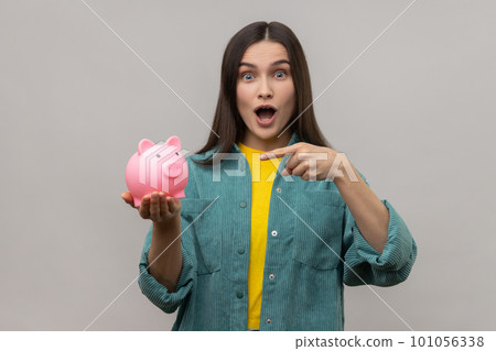 Portrait of amazed woman with dark hair standing pointing at piggy bank in her hand, advantageous bank offer, wearing casual style jacket. Indoor studio shot isolated on gray background. 101056338
