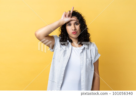Portrait of depressed woman with dark wavy hair holding hand near forehead showing loser gesture with fingers, having problems, looks unhappy. Indoor studio shot isolated on yellow background. Portrait of depressed woman with dark wavy hair holding hand near forehead showing loser gesture with fingers, having problems, looks unhappy. Indoor studio shot isolated on yellow background. 101056340