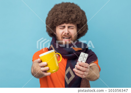 Portrait of man with Afro hairstyle wearing orange T-shirt and wrapped in scarf, treating with pills and warm tea, being upset, feeling unwell. Indoor studio shot isolated on blue background. 101056347