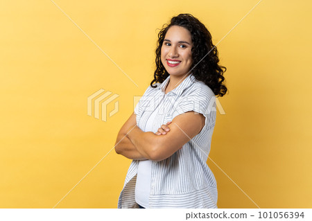 Portrait of satisfied delighted happy woman with dark wavy hair standing with folded hands and looking at camera with toothy smile. Indoor studio shot isolated on yellow background. 101056394