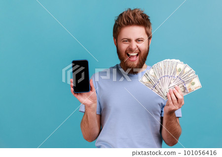 Portrait of extremely happy handsome bearded man holding dollar banknotes and cell phone with empty screen, online betting, winning. Indoor studio shot isolated on blue background. 101056416