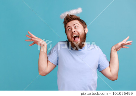 Portrait of handsome funny bearded angelic man with holy nimbus on head standing with raised hands and looking away with open mouth. Indoor studio shot isolated on blue background. Portrait of handsome funny bearded angelic man with holy nimbus on head standing with raised hands and looking away with open mouth. Indoor studio shot isolated on blue background. 101056499