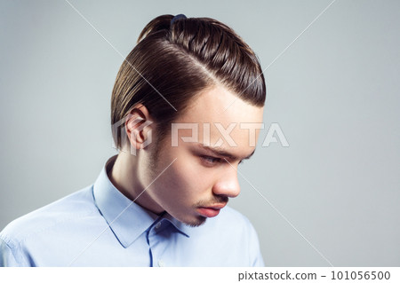 Side view portrait of young adult brunette man with mustache and beard with top knot hairstyle, looking away, wearing blue shirt. Indoor studio shot isolated on gray background. Side view portrait of young adult brunette man with mustache and beard with top knot hairstyle, looking away, wearing blue shirt. Indoor studio shot isolated on gray background. 101056500
