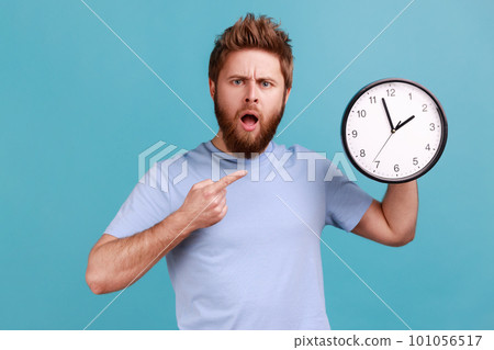 Portrait of shocked astonished surprised handsome bearded man pointing at wall clock, deadline, having puzzled facial expression. Indoor studio shot isolated on blue background. Portrait of shocked astonished surprised handsome bearded man pointing at wall clock, deadline, having puzzled facial expression. Indoor studio shot isolated on blue background. 101056517
