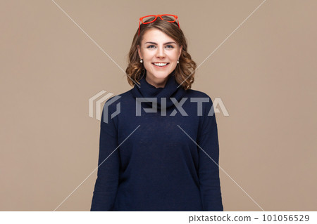 Portrait of positive smiling happy woman with wavy hair in red red glasses on her head looking at camera with satisfied expression. Indoor studio shot isolated on light brown background. 101056529