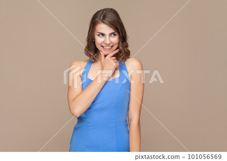 Portrait of woman with wavy hair having cunning look, holding hand on chin having some tricky plans in her mind, wearing blue dress. Indoor studio shot isolated on light brown background. 101056569