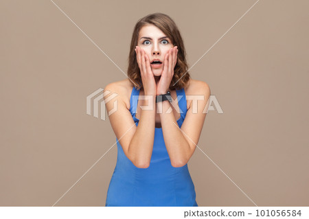 Portrait of shocked surprised beautiful woman with wavy hair standing with palms on her cheeks, sees something astonished, wearing blue dress. Indoor studio shot isolated on light brown background. 101056584