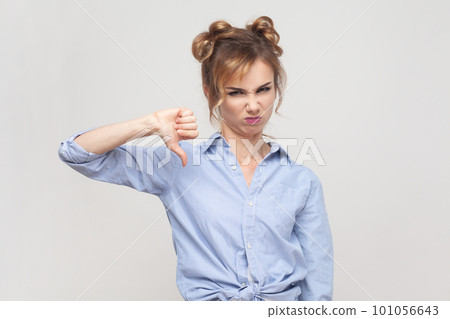 Portrait of disappointed blonde woman shows disapproval sign, keeps thumb down, expresses dislike, frowning face in discontent, wearing blue shirt. Indoor studio shot isolated on gray background. 101056643
