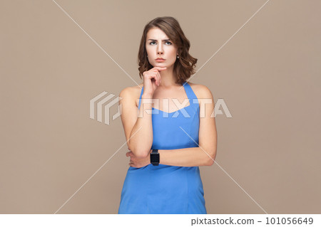 Portrait of pensive thoughtful beautiful woman with wavy hair standing and holding chin, thinking about future plans, wearing blue dress. Indoor studio shot isolated on light brown background. 101056649