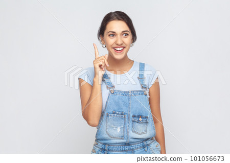 Portrait of inspired excited woman wearing denim overalls pointing finger up and looking amazed about sudden genius idea, got solution. Indoor studio shot isolated on gray background. Portrait of inspired excited woman wearing denim overalls pointing finger up and looking amazed about sudden genius idea, got solution. Indoor studio shot isolated on gray background. 101056673