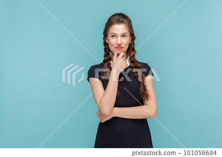 Portrait of cunning woman with braids standing holding chin pondering, thinking about devil plan or prank, wearing black dress. woman Indoor studio shot isolated on blue background. Portrait of cunning woman with braids standing holding chin pondering, thinking about devil plan or prank, wearing black dress. woman Indoor studio shot isolated on blue background. 101056684