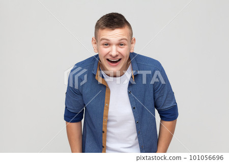 Portrait of overjoyed extremely happy handsome teenager boy wearing blue shirt hunching from laughter, laughing out loud, crazy face. Indoor studio shot isolated on gray background. Portrait of overjoyed extremely happy handsome teenager boy wearing blue shirt hunching from laughter, laughing out loud, crazy face. Indoor studio shot isolated on gray background. 101056696