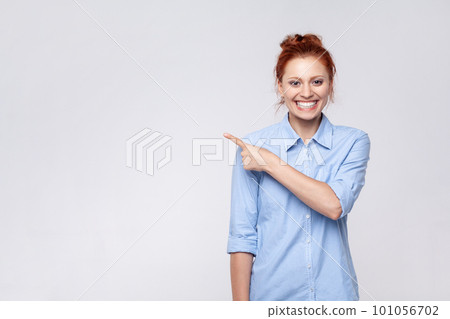 Portrait of ginger woman wearing blue shirt looking at camera and pointing away, presenting copy space for advertisement or promotion. Indoor studio shot isolated on gray background. Portrait of ginger woman wearing blue shirt looking at camera and pointing away, presenting copy space for advertisement or promotion. Indoor studio shot isolated on gray background. 101056702