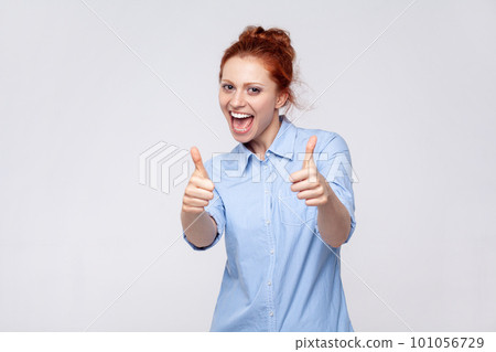 Portrait of joyful cheerful delighted young adult ginger woman wearing blue shirt showing thumbs up, enjoying service, recommend. Indoor studio shot isolated on gray background. 101056729