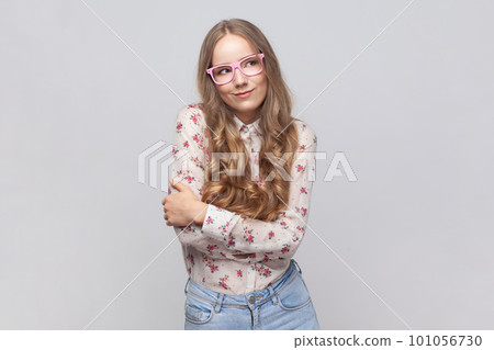Portrait of woman in glasses with wavy blond hair standing hugging herself and feeling good, appreciating her appearance, self esteem concept. Indoor studio shot isolated on gray background. Portrait of woman in glasses with wavy blond hair standing hugging herself and feeling good, appreciating her appearance, self esteem concept. Indoor studio shot isolated on gray background. 101056730