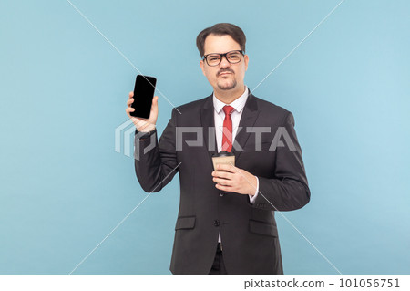 Portrait of sad man with mustache showing smart phone with blank screen, frowning face, drinking coffee to go, wearing black suit with red tie. Indoor studio shot isolated on light blue background. 101056751