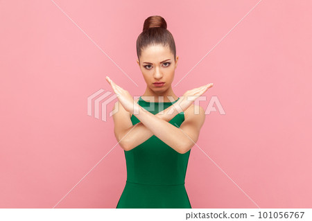 Portrait of attractive strict serious woman with bun hairstyle standing and showing x sign, no way gesture, prohibition, wearing green dress. Indoor studio shot isolated on pink background. 101056767