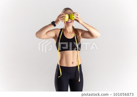 Portrait of funny athletic blonde woman standing with green apples in hands, covering eyes with fresh fruit, wearing black fitness clothing. Indoor studio shot isolated on gray background. 101056873