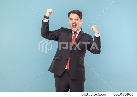 Portrait of excited extremely happy man standing with clenched fists, celebrating his victory, screaming hurray, wearing black suit with red tie. Indoor studio shot isolated on light blue background. Portrait of excited extremely happy man standing with clenched fists, celebrating his victory, screaming hurray, wearing black suit with red tie. Indoor studio shot isolated on light blue background. 101056903