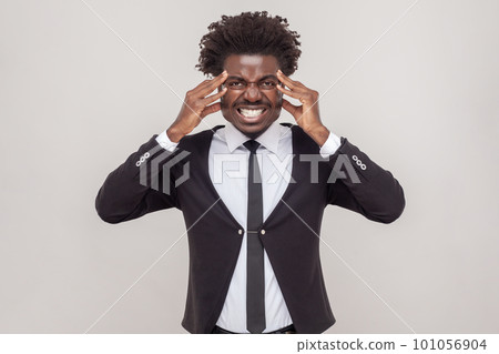 Portrait of unhappy man with petit goatee suffers from unbearable headache, keeps hands on temples, needs painkillers, wearing white shirt and tuxedo. Indoor studio shot isolated on gray background. 101056904