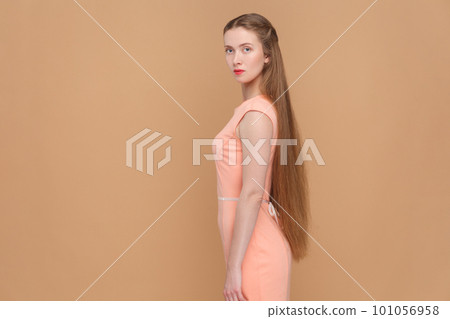 Side view portrait of serious strict woman with long hair looking at camera with bossy expression, being in bad mood, wearing elegant dress. Indoor studio shot isolated on brown background. 101056958