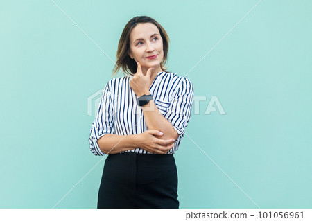 Portrait of pensive attractive middle aged woman wearing striped shirt standing holding chin, thinking about future plans. Indoor studio shot isolated on light blue background. 101056961