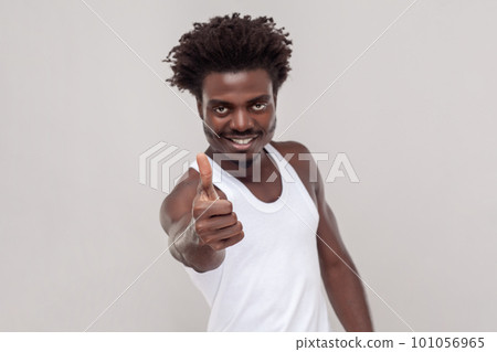 Very good. Pleased nice looking man with Afro hairstyle showing thumb up, gives positive feedback, demonstrates approval, wearing white T-shirt. Indoor studio shot isolated on gray background. 101056965
