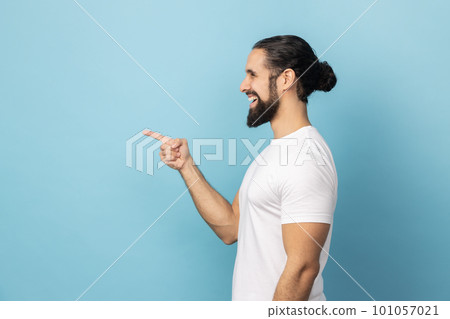 Side view of smiling man with beard in white T-shirt pointing finger aside, showing copy space for commercial text, blank wall with idea presentation. Indoor studio shot isolated on blue background. Side view of smiling man with beard in white T-shirt pointing finger aside, showing copy space for commercial text, blank wall with idea presentation. Indoor studio shot isolated on blue background. 101057021