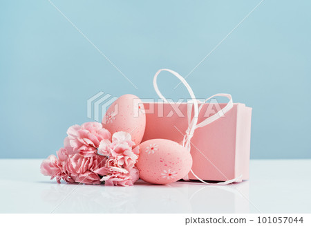 Two Easter eggs with pink gift box and tender carnations spring flowers on white table blue background. Side view. Happy Easter in pink. Copy space 101057044