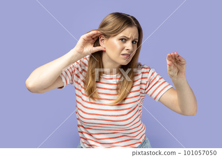 Portrait of blond woman wearing striped T-shirt holding hand near ear trying to listen quiet conversation, overhearing gossip, showing blah gesture. Indoor studio shot isolated on purple background. 101057050
