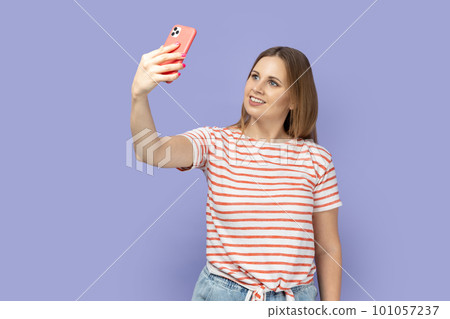 Portrait of smiling blond woman in striped T-shirt standing with phone and having video call or broadcasting livestream, looking smiling at screen. Indoor studio shot isolated on purple background. 101057237