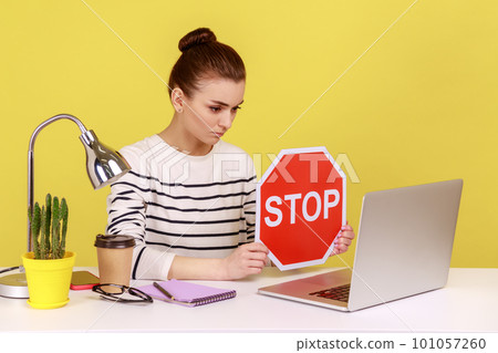 Woman manager wearing striped shirt showing stop red sign to laptop screen, avoiding conflicts, afraid of workplace bullying. Indoor studio studio shot isolated on yellow background. Woman manager wearing striped shirt showing stop red sign to laptop screen, avoiding conflicts, afraid of workplace bullying. Indoor studio studio shot isolated on yellow background. 101057260