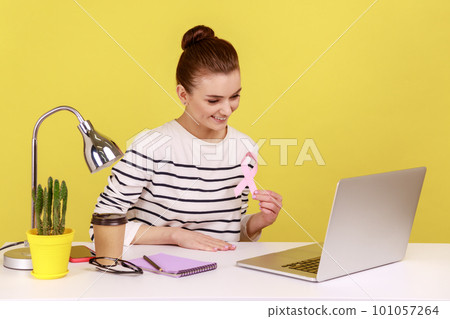 Delighted woman volunteer sitting at workplace showing pink ribbon to laptop screen, symbol of breast cancer awareness, female health. Indoor studio studio shot isolated on yellow background. 101057264