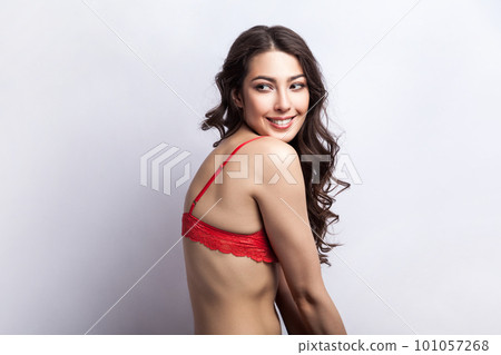 Side view portrait of smiling satisfied shy woman sensuality model with curly hair looking away with smile, hiding her red lingerie. Indoor studio shot isolated on gray background. 101057268