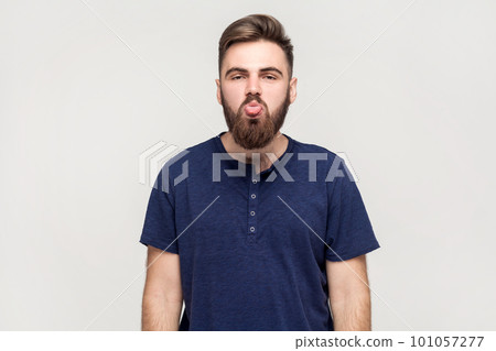 Portrait of man with beard wearing dark blue T-shirt showing out tongue and looking at camera with naughty disobedient grimace, making face. Indoor shot isolated on gray background. 101057277